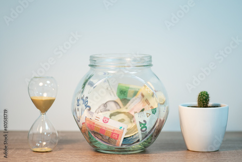 Glass jar with Ukrainian money inside, sandglass and cactus ner it on the table, close up. Concept of savings, financial planning, wealth and poorness