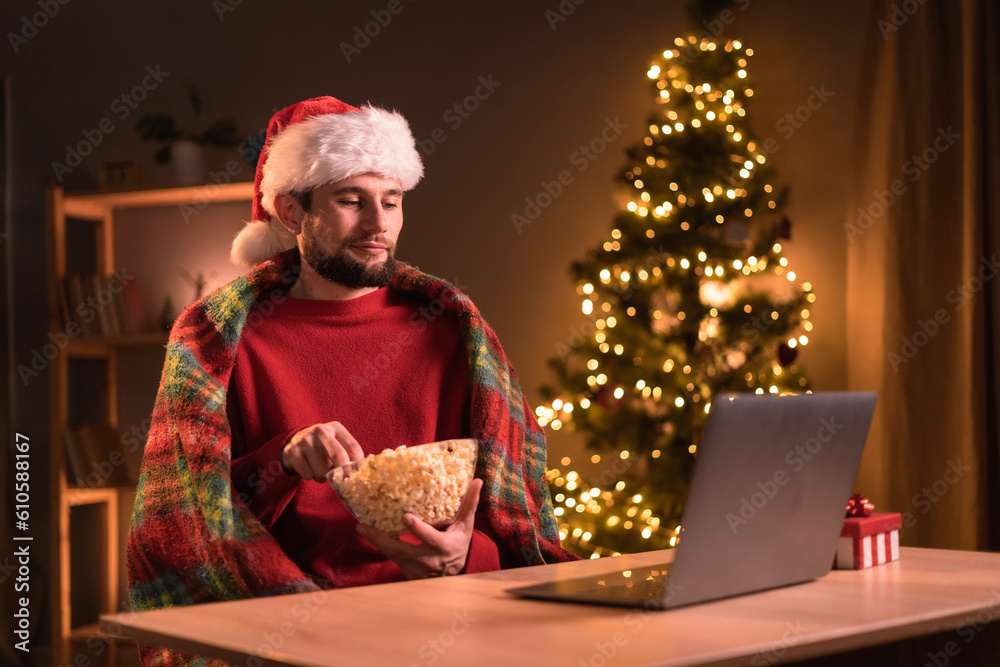 Christmas eve. Alone young man watching movie using laptop and eating popcorn. Home cinema.