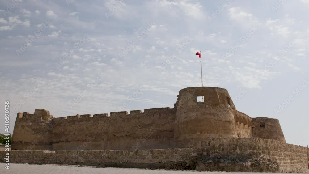 A wide-shot view of Arad Fort in Bahrain. Three towers are on show between stone walls. The flag of Bahrain flies above one of the towers against a blue sky.