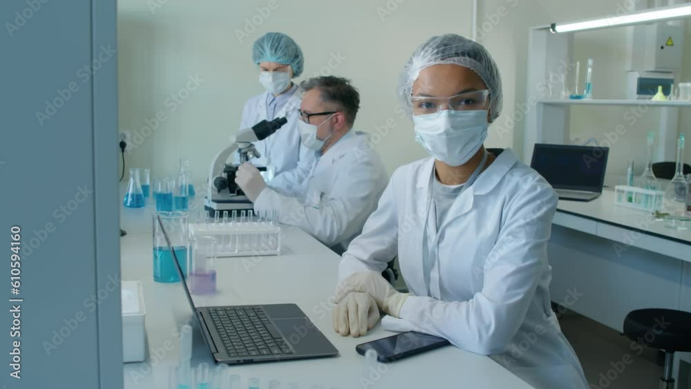 Portrait of Biracial female scientist wearing goggles, mask and medical gown sitting at desk in laboratory at daytime while two colleagues in background working with microscope Portrait of Biracial female scientist wearing goggles, mask and medical gown sitting at desk in laboratory at daytime while two colleagues in background working with microscope