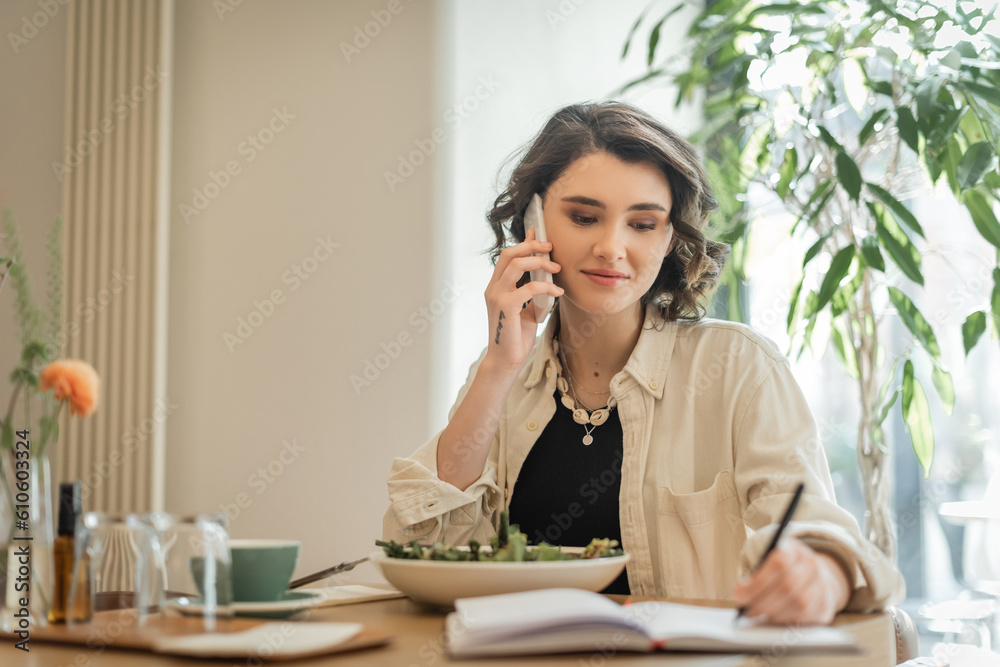 smiling woman with wavy brunette hair and tattoo talking on mobile phone and writing in notebook near vegetable salad and cup of aromatic coffee in lobby cafe of modern hotel, freelance lifestyle smiling woman with wavy brunette hair and tattoo talking on mobile phone and writing in notebook near vegetable salad and cup of aromatic coffee in lobby cafe of modern hotel, freelance lifestyle