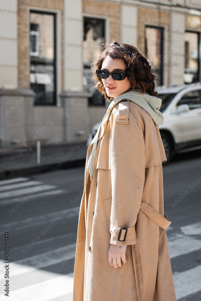 Obraz premium attractive woman with wavy brunette hair walking in dark stylish sunglasses, grey hoodie and beige trench coat while looking at camera on blurred street in European city, urban fashion