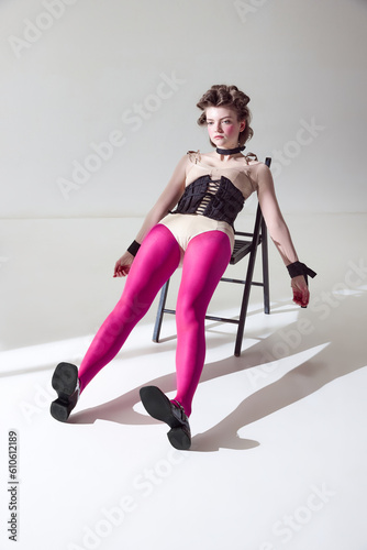 Portrait of young girl posing on chair in black corset and pink tights against grey studio background. Weirdness and uniqueness