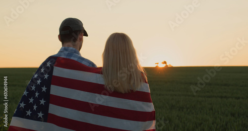 Two farmers watch how a tractor works in the field. Standing side by side at sunset with an American flag on their shoulders.