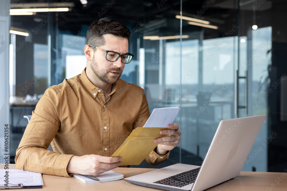 Fototapeta premium A serious and worried young businessman working in the office holds a letter, documents. got bad news. Saddenedly reads a message, a contract, an agreement