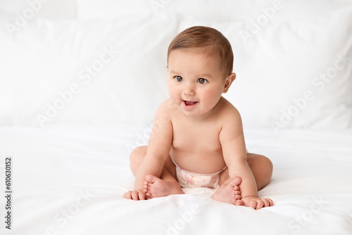 Baby girl wearing cloth diaper sitting on white bed leaning on hands with funny expression photo