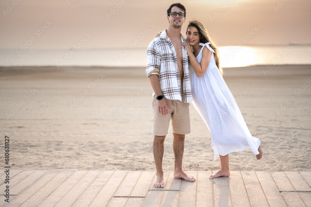 Full length portrait of pregnant woman in white dress leaning over her husband shoulders while standing and posing together on wooden walkway on the beach. Full length portrait of pregnant woman in white dress leaning over her husband shoulders while standing and posing together on wooden walkway on the beach.