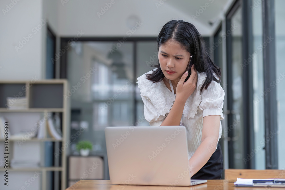 Portrait of smiling young Asian female manager working remotely, holding paper and reading financial document, talking on mobile phone standing at desk at home office