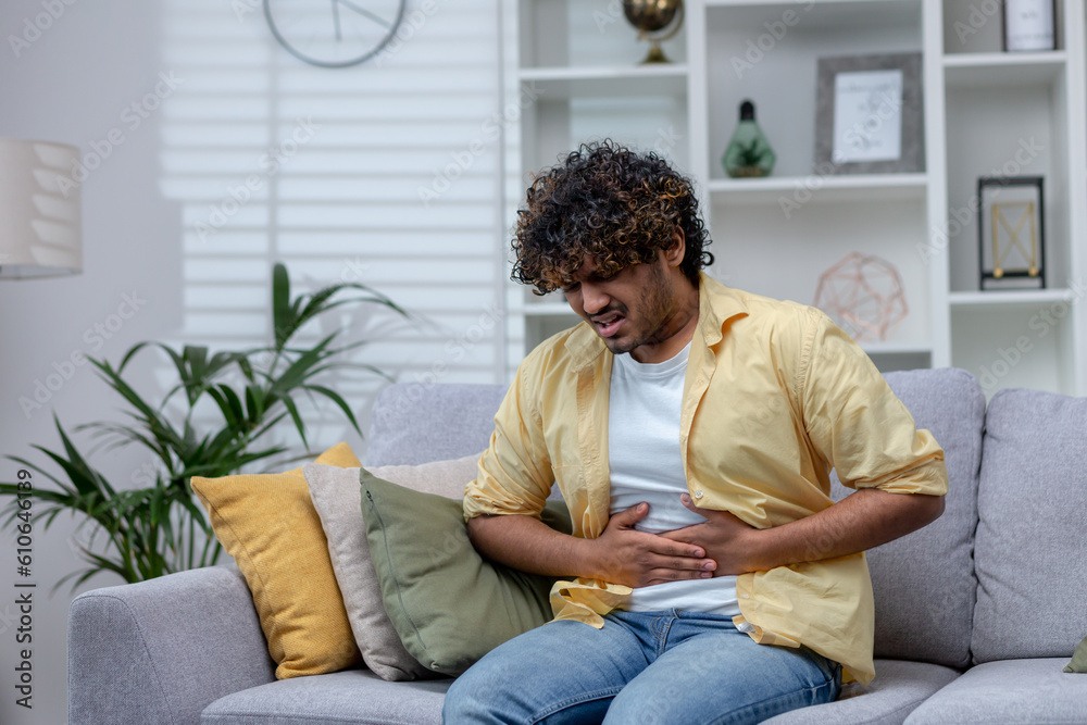 Young man sick at home sitting on sofa, Latin American has severe stomach pain and nausea, poisoned by poor quality food. Young man sick at home sitting on sofa, Latin American has severe stomach pain and nausea, poisoned by poor quality food.