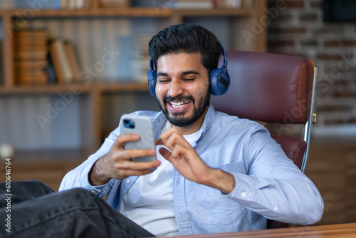 Smiling indian business man office worker, male executive manager wearing headphones sitting at work using phone listening to podcast or remote video chatting by virtual call.
