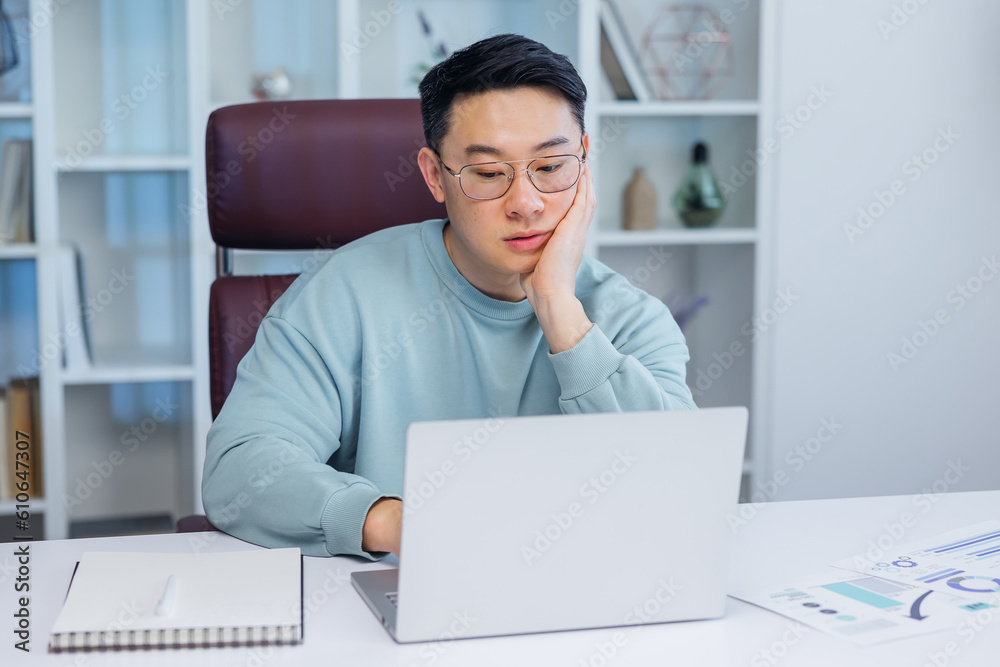 Bored young Asian guy sitting at desk using laptop, thinking about something during online lesson or remote work at home office. Tired man having dull distant job, feeling lazy Bored young Asian guy sitting at desk using laptop, thinking about something during online lesson or remote work at home office. Tired man having dull distant job, feeling lazy
