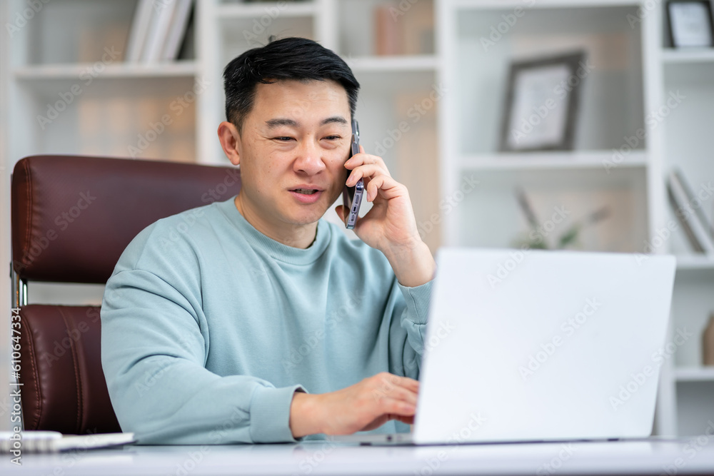 Handsome asian manager sitting at office desk in front of laptop hold mobile phone make pleasant business or informal call. Handsome asian manager sitting at office desk in front of laptop hold mobile phone make pleasant business or informal call.