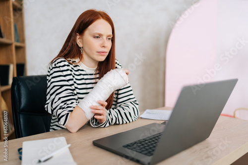 Tired injured young woman with broken right hand wrapped in white gypsum bandage focused looking on laptop computer screen sitting at table. Sick businesswoman working remotely from home office.