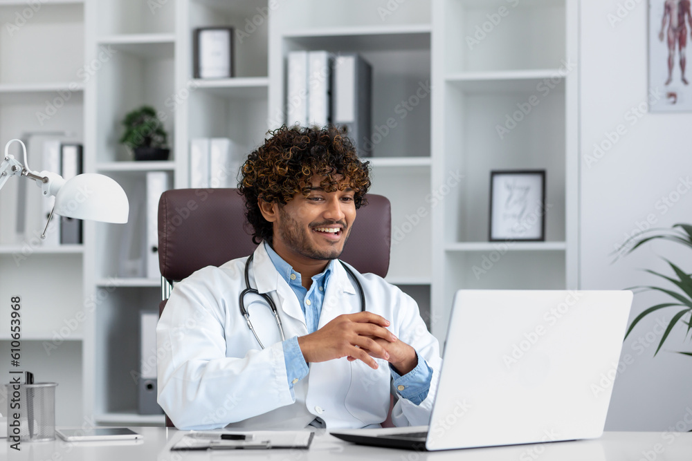 Young successful doctor working inside hospital office, man smiling and looking at laptop screen, hispanic man in medical gown is satisfied with patient's treatment achievement results.