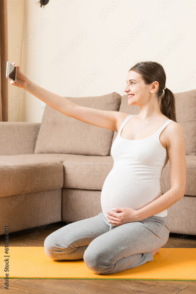 Pregnant female holding smartphone while sitting on exercise yoga mat at home. Fitness or yoga training online at coronavirus time Pregnant female holding smartphone while sitting on exercise yoga mat at home. Fitness or yoga training online at coronavirus time