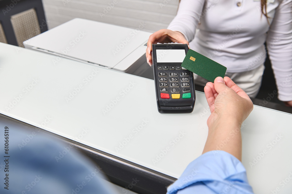 Hand of customer paying with contactless credit card with NFC technology. Female administrator with credit card reader machine at reception counter with female holding blank bank card. Selective focus Hand of customer paying with contactless credit card with NFC technology. Female administrator with credit card reader machine at reception counter with female holding blank bank card. Selective focus
