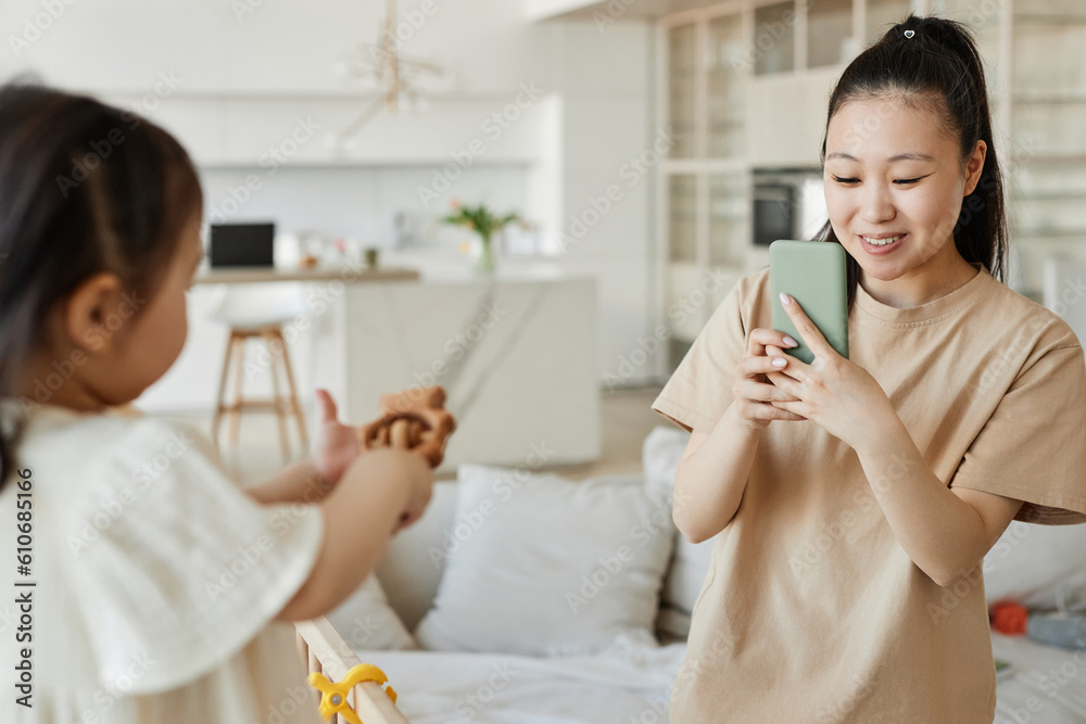 Happy Asian mom making photo of her kid on smartphone while they playing in the room Happy Asian mom making photo of her kid on smartphone while they playing in the room