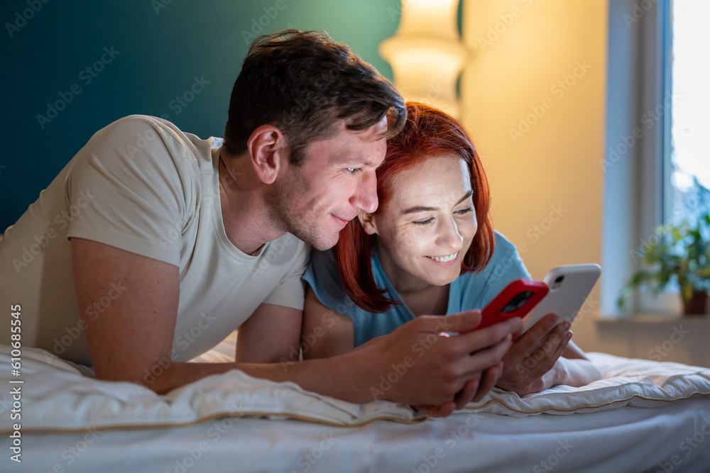 Couple looking phone while lying on bed. Girl showing boyfriend funny videos on phone. Happy