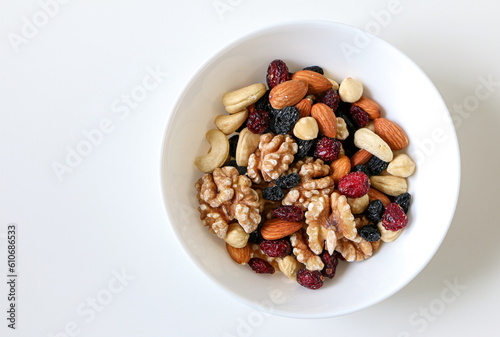 Top view of a white bowl of mixed nuts and dried fruits consisting of almonds, walnuts, hazelnuts, cashew nuts, raisins and cranberries, on right side of white table, copy space on left