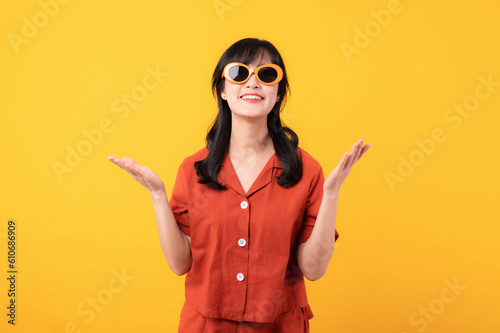 Portrait young beautiful asian woman happy smile dressed in orange clothes and sunglasses showing hand gesture, arm compare two variants demonstrate empty space isolated on yellow studio background. © Jirawatfoto