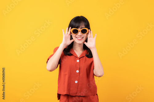Portrait young beautiful asian woman happy smile dressed in orange clothes and sunglasses looking surprised, reacting amazed, raising eyebrows impressed isolated on yellow studio background.