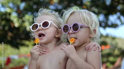 Slow motion close-up naked kids sisters in park on picnic outside licking lollipops on stick. Concept of happy childhood with variety of taste impressions. Naked kids eating lollipops slow motion photo