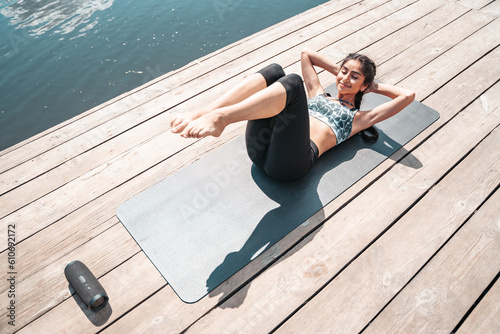 Young Indian woman doing abs exercise on wooden pier by the river enjoying summer morning fresh air