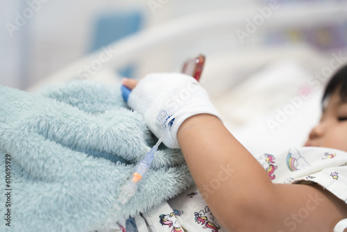 child is receiving medication through intravenous fluid therapy in hospital bed, focus on drip, and using smart phone