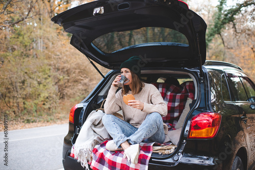 Young woman wear knitted clothes drinking hot tea and resting in car at road outdoor over autumn nature at background. Travel lifestyle. © morrowlight