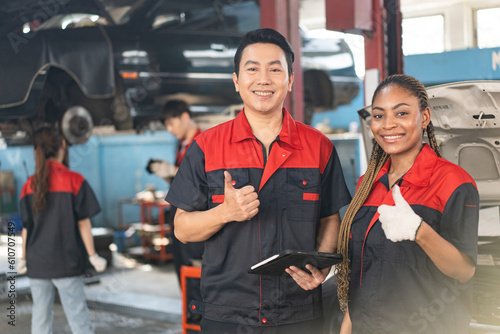Selective focus of mid-adult Asian male and young African female mechanics in uniforms, standing holding tablet, showing thumbs up and smiling at camera with mechanics repairing car in the background.