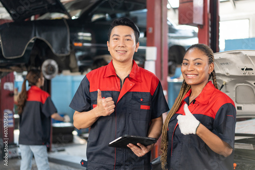 Selective focus of mid-adult Asian male and a young African female mechanics in uniforms, standing holding a tablet, showing thumbs up and smiling at the camera with blurred garage in the background.