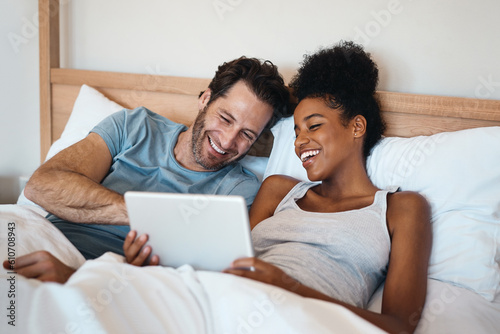 Happy couple, tablet and laughing in bed for funny entertainment, joke or morning in relax together at home. Interracial man and woman person with laugh on technology for social media meme in bedroom