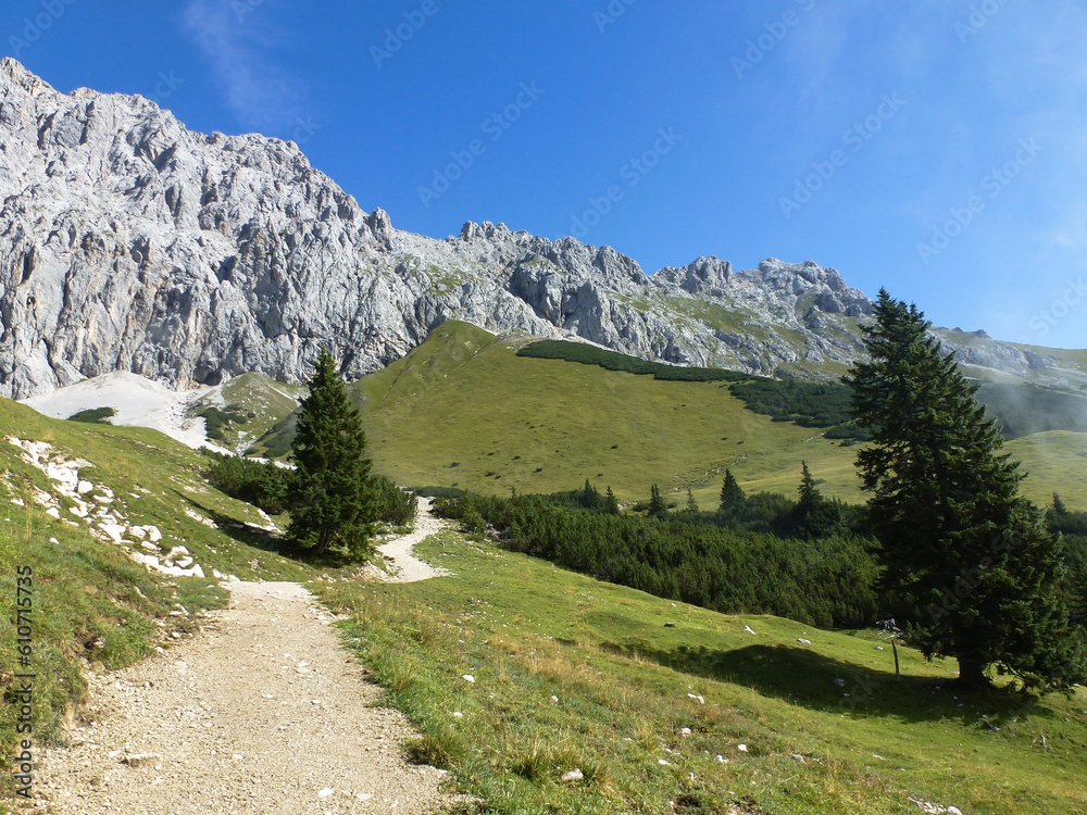 A rock climbing trail stretches across the green plateau to the rocky mountains. Summer landscape of mountain nature