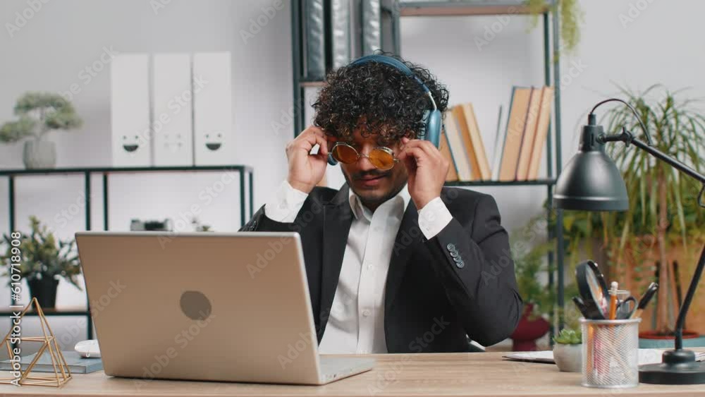 Happy relaxed overjoyed indian businessman working on laptop computer at home office wearing headphones listening favorite energetic disco music and dancing. Freelancer man relaxing, taking a break Happy relaxed overjoyed indian businessman working on laptop computer at home office wearing headphones listening favorite energetic disco music and dancing. Freelancer man relaxing, taking a break
