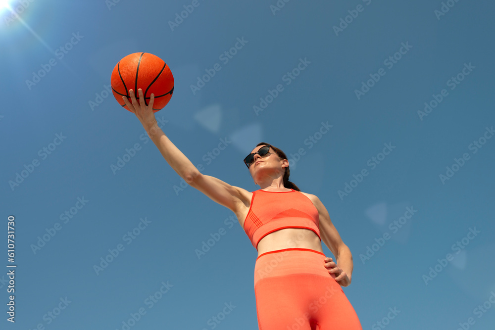 Sporty, fit woman holding a basketball, blue sky background.