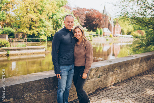 middle aged couple standing in front of a bridge over the water in an english countryside in europe, reflections in the water, greenery, fall, autumn