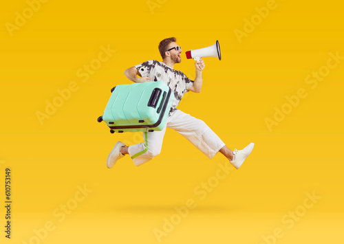 Portrait of a funny young man tourist wearing casual clothes with suitcase screaming in mouthpiece on studio yellow background and jumping. Vacation trip and summer holiday concept.