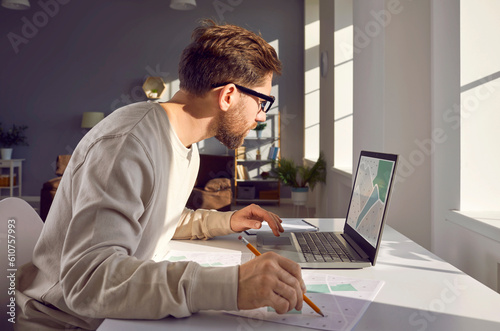 Young man sitting at his desk or table, working with cadastral city maps, using a modern laptop PC, looking at the lake or pond location on the computer screen, and taking notes on printed paper maps