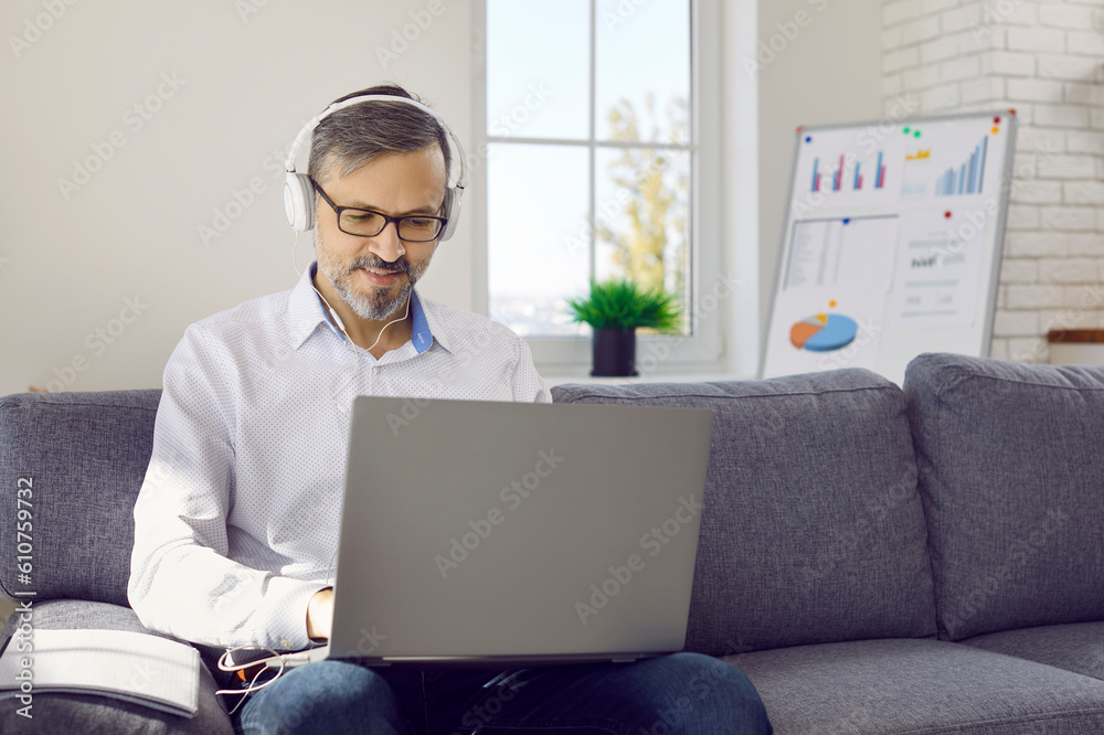 Mature man studying online. Adult student in headphones and glasses sitting on sofa at home, typing on laptop computer, doing business marketing training course, searching for new useful information