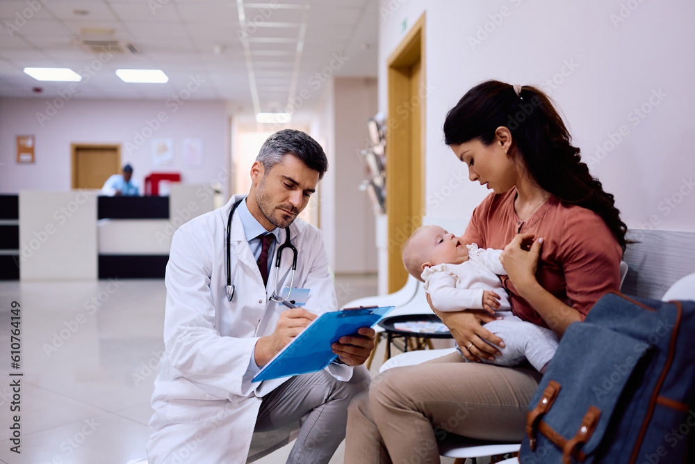 Male doctor filling data in medical record while talking to mother with baby in waiting room at clinic.