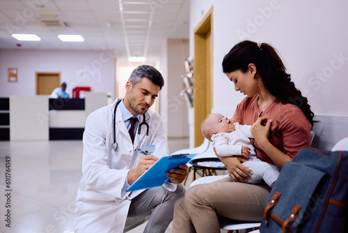 Male doctor filling data in medical record while talking to mother with baby in waiting room at clinic.