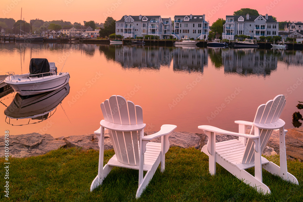Sunrise, two white Adirondack chairs on the beach, a moored boat, and  wildfire smoke over the Mystic River marina village in Connecticut