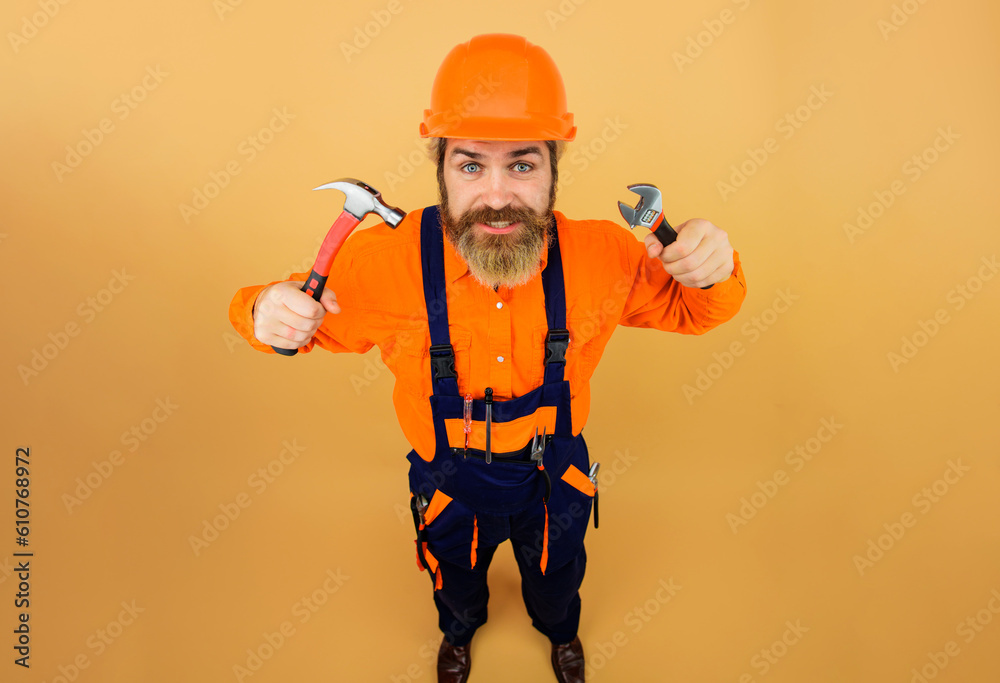 Bearded builder in safety helmet and overalls with hammer and adjustable spanner. Construction worker in hardhat with tools for repair. Repairman, craftsman or foreman with pipe wrench and hammer.