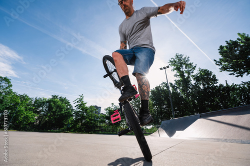 Low angle view of a middle-aged tattooed man performing tricks on his bmx bike in a skate park.