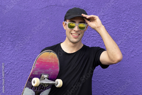 Young caucasian skateboarder smiling happy holding skateboard over violet wall. Street photo.