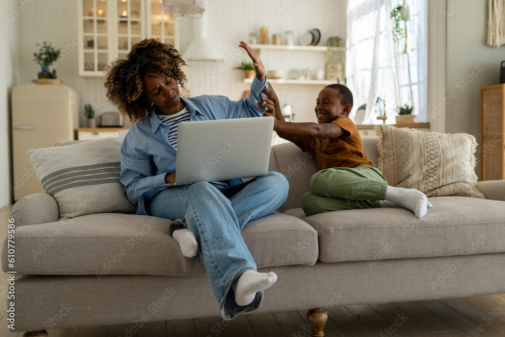 Small child disturbing work-at-home mom. Displeased young african american mother ignoring son while job remotely on laptop from home. Parent freelancer. No nanny forchild when parents working