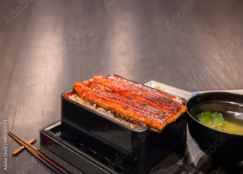 Fillets of japanese unagi eel  grilled in a kabayaki style topped on steamed white rice in a lackered jubako food box aside a cup of misoshiru soup and chopsticks on chopstick a holder on a table. photo