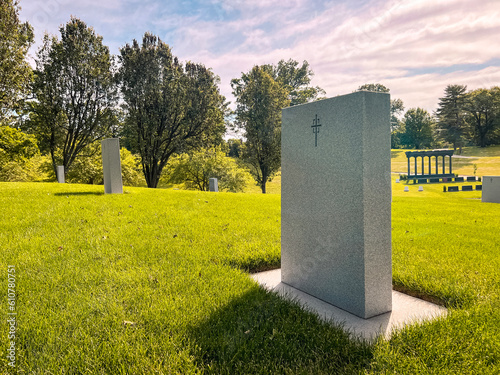 Focus on a single empty tombstone in a row of unmarked gravesites. The front of the stone is blank, except for a small engraved cross at the top of the headstone. Situated in a lush green cemetery.  photo