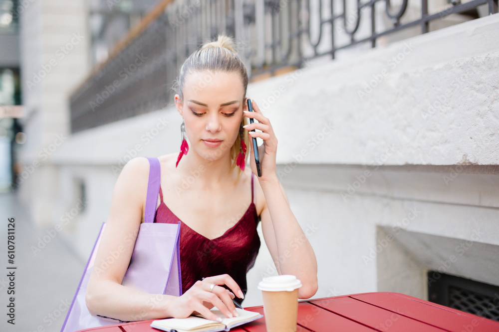 Fototapeta premium Young woman sitting at table writing notes using her cell phone