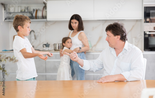 At home in kitchen, parents scold son for imprudent act committed. Sulky schoolboy boy stands with arms crossed in front of, parents during serious conversation with boy Mom hugs daughter